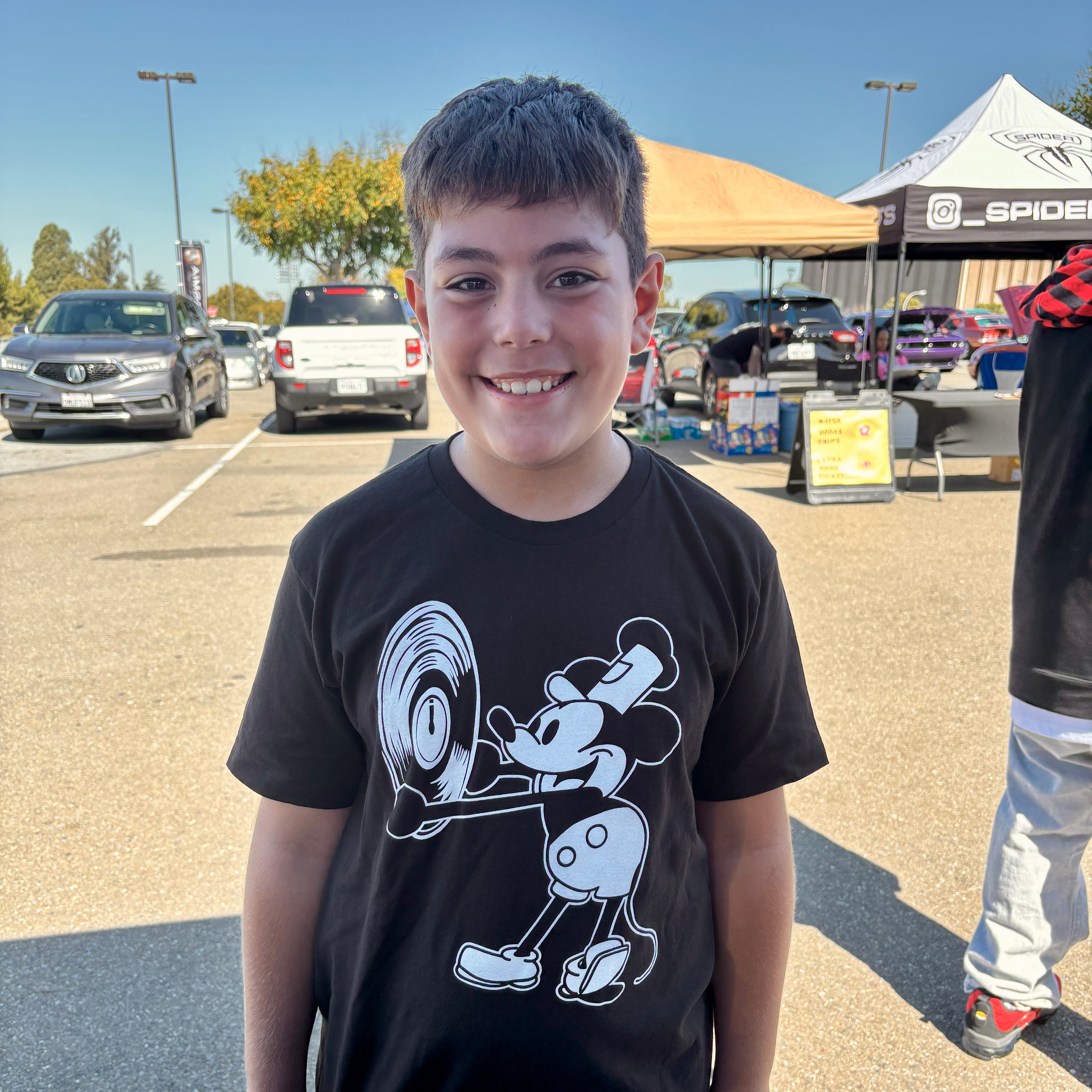 Young boy wearing a black t-shirt mickey shirt holding vinyl with a graphic design, standing in a parking lot at car show