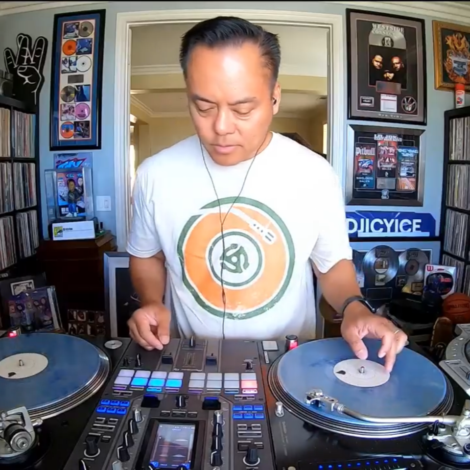 DJ operating turntables and mixer in a room filled with records and DJ equipment.